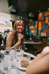 A happy young woman is captured mid-laughter as she smokes electronic cigarette in a lively, colorful cafe environment, suggesting a light-hearted conversation.