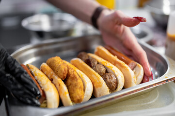 Close-up view hands preparing hot dogs with various toppings on metal tray, showcasing process of...