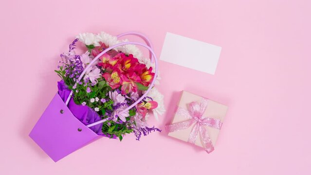 Bouquet of fresh flowers in purple bag along with white card and gift box appear on pink background. Greeting card for birthday, mother's day, women's day or other occasion. Stop motion animation. Cop