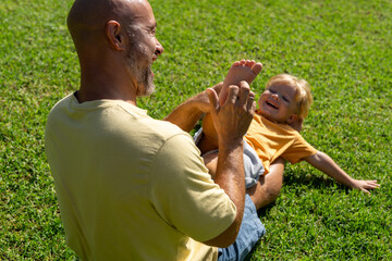 family and love concept - young father tickling his little son in park. dad tickles his son's legs on the grass. dad with little son having fun on the green lawn - obrazy, fototapety, plakaty
