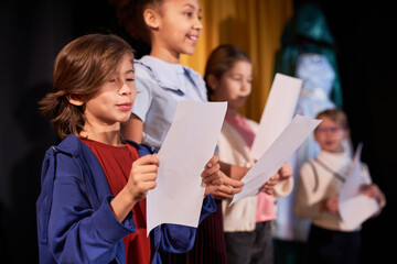 Side view portrait of young boy rehearsing lines standing on stage in school theater in row with...