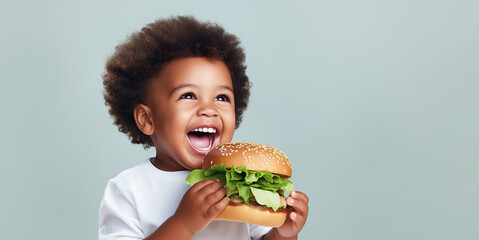 Happy African American child bites a delicious hamburger with lettuce. A cheerful little boy in a white T-shirt holds a hamburger in his hands on a gray background. Banner with place for text - obrazy, fototapety, plakaty