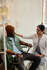 Vertical portrait of caring young nurse talking to patient receiving IV treatment in clinic