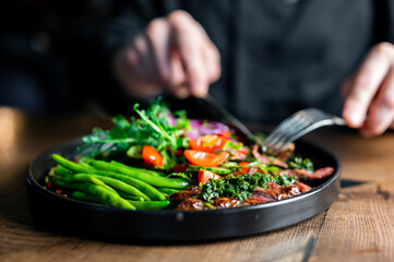A person enjoys a vibrant, healthy meal of green beans, salad, and grilled meat on a dark plate....