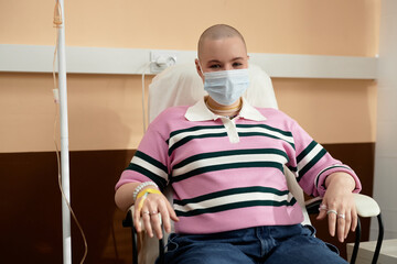 Front view portrait of bald young woman wearing mask getting IV drip treatment in clinic and...