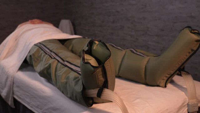 Man lying on couch visits physiotherapy clinic to undergo body pressotherapy session through pneumatic suit to improve blood circulation and lymphatic system.