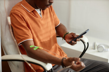 Side view closeup of African American adult man using smartphone during IV therapy procedure in...