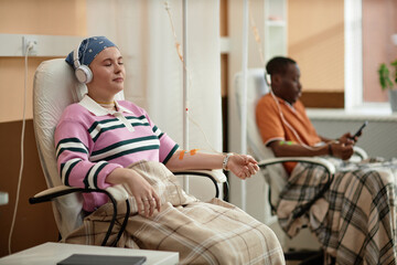 Portrait of young woman listening to music while getting chemotherapy treatment in clinic sitting in chair with IV drip copy space - obrazy, fototapety, plakaty