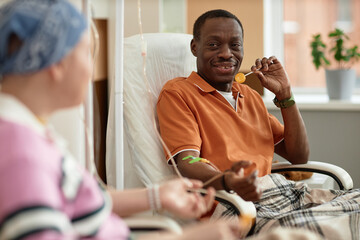 Portrait of smiling African American man talking to another patient and holding loli during chemotherapy treatment in clinic - obrazy, fototapety, plakaty