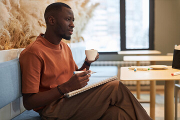 Side view portrait of Black young man as artist drawing sketches in cozy coffee shop and enjoying...