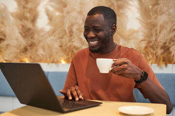 Portrait of smiling Black man in online meeting via laptop working in cozy coffee shop copy space