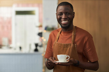 Waist up portrait of smiling African American man as cafe worker holding coffee cup and wearing apron, copy space - obrazy, fototapety, plakaty