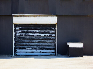 Closed metal shutter on black wall. Small shop's entrance shutter door in a beautiful black background wall. Industrial background pattern.