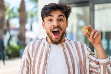 Handsome Arab man holding a Bitcoin at outdoors with surprise and shocked facial expression