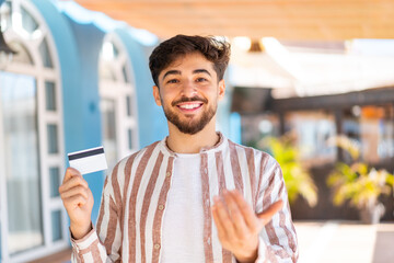 Handsome Arab man holding a credit card at outdoors inviting to come with hand. Happy that you came