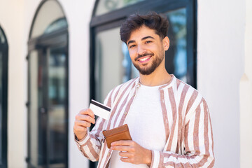 Handsome Arab man at outdoors holding wallet and credit card with happy expression