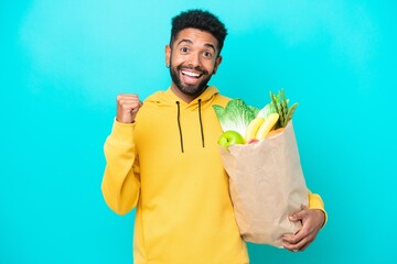 Young Brazilian man taking a bag of takeaway food isolated on blue background celebrating a victory...