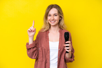 Young singer caucasian woman picking up a microphone isolated on yellow background showing and lifting a finger in sign of the best