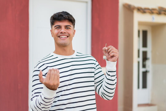 Young hispanic man holding home keys at outdoors inviting to come with hand. Happy that you came