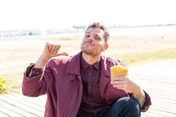 Young caucasian man holding fried chips at outdoors proud and self-satisfied
