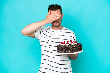 Young caucasian man holding birthday cake isolated on blue background covering eyes by hands. Do...