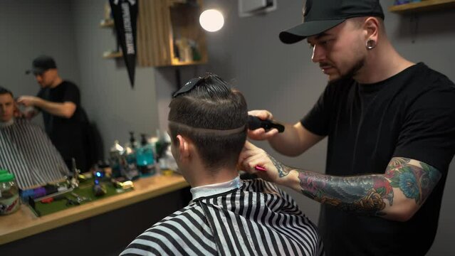 A barber man with a tattoo on his arm makes a haircut to a brunette guy in a barbershop, cuts his hair with a clipper close-up