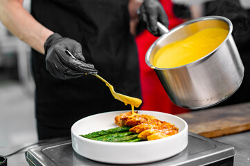 chef in black attire garnishing a dish of grilled chicken and green vegetables with yellow sauce from a steel pot. The plate is neatly arranged on a white surface, emphasizing the vcolors of the food