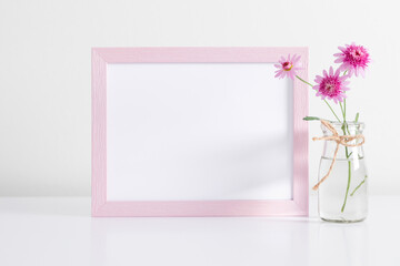 Blank frame mockup with pink flowers on table on white wall background. Empty frame mockup.
