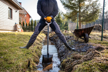 Man in protective gear tirelessly working to dredge a muddy trench with a shovel and wheelbarrow in...