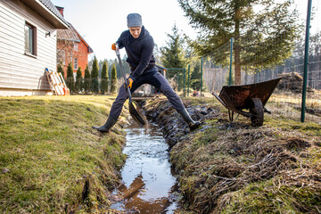 Man in workwear clearing a waterlogged garden path in early spring with a shovel and wheelbarrow...