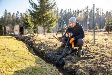Man pausing from outdoor work to check his phone while kneeling by a newly dug drainage ditch in a...