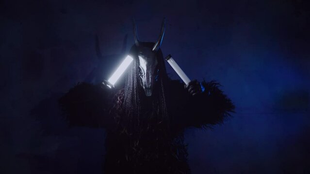 The devil monster dances with RGB sticks in his hands. A mystical man in a scary mask in a dark studio. The man is dressed in a black coat and a mask with horns on his head. Alien from space. Horror, 