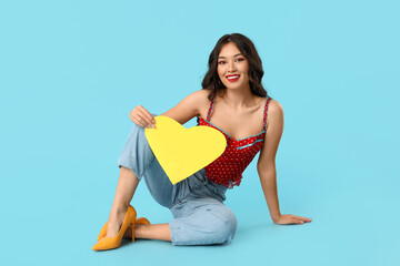 Beautiful young happy Asian woman with paper heart sitting on blue background. Valentine's Day...