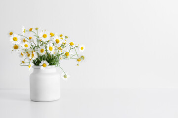 White chamomile flowers in a white pot. Bouquet of small fresh daisy flowers in  vase on background of white wall. Elegant floral arrangement with wildflowers. Copy space, front view