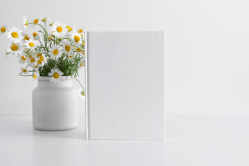 White book mockup, diary, white table, white chamomile flowers in vase. Front view. Place for text,...
