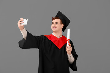 Male graduating student with diploma taking selfie on grey background