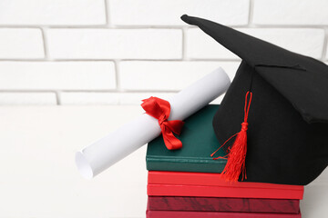 Mortar board with diploma and books on table near white brick wall