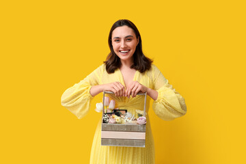 Happy young woman holding Easter basket with decorative cosmetics on yellow background