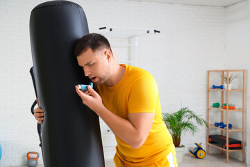 Sporty young man using inhaler near punching bag in gym