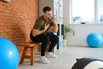 Sporty young man using inhaler on bench in gym