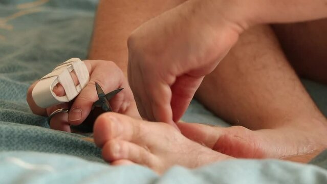 A man cuts his toenails with a broken toe on the bed at home, doing a pedicure himself