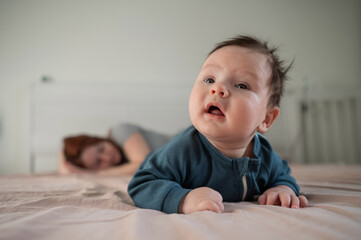 Fototapeta premium A three-month-old boy lies on his stomach on the bed and his mother sleeps behind him. Postpartum depression.