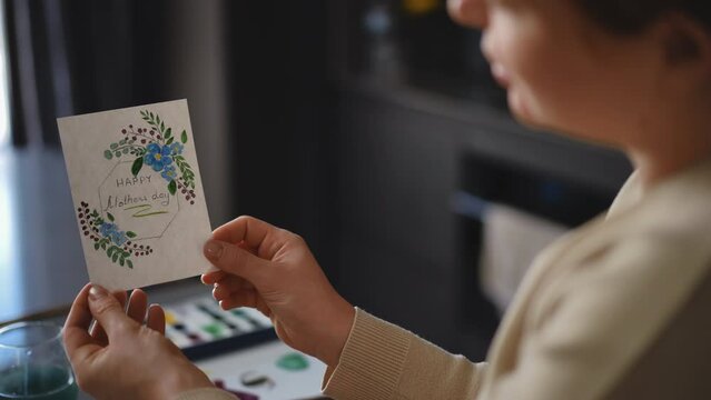 A woman holds a Mother's Day greeting card, which she painted with watercolors, while sitting at a table in the living room. DIY greeting card