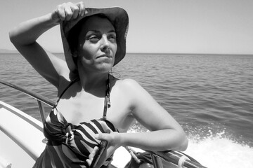 Black and white of a woman on a boat, wearing a swimsuit with a hat and holding a smartphone.