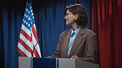 Medium shot of mature Caucasian female politician in glasses, suit jacket and blouse, walking on stage, standing behind rostrum, looking around audience, smiling, waving hand, greeting electorateMediu