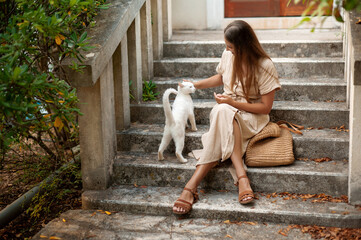 Young pleasant lady sitting on street staircase with crossed legs and petting white homeless cat....