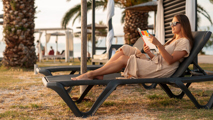 Side view of attractive caucasian lady wearing beige sundress and black sunglasses lying on deck chair and reading scientific book. Long-haired female spending pastime among palm trees on beach.
