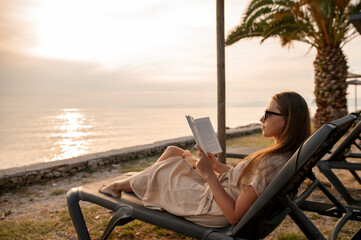 Back angle view of young beautiful woman lying on deck chair in front of seacoast and reading interesting book. Charming brunette dressed in light beige dress relaxing on beach during sunset.