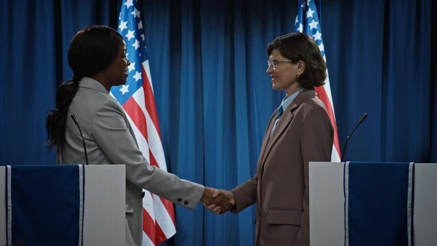 Medium shot of African American and Caucasian female Senate candidates standing on stage, waving to public, then shaking hands, greeting each other and wishing luck, before beginning debate