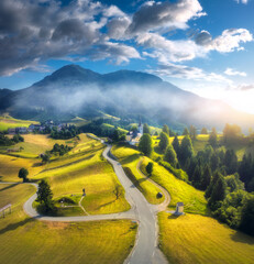 Aerial view of country road in green meadows at sunset in summer. Top drone view of rural road, alpine mountains in low clouds. Colorful landscape with hills, fields, green grass, blue sky. Slovenia